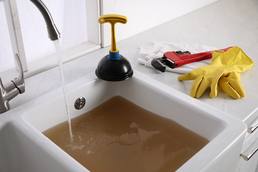 A clogged sink filled with dirty water; there is a pair of gloves, a tool, and a plunger on the countertop beside it.